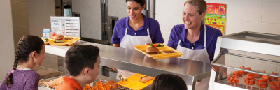 Two lunch monitors serving lunches to children in school in a cafeteria line.